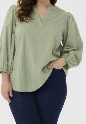 Woman wearing light green striped blouse with gathered sleeves and dark blue jeans, hand in pocket, against a plain background.