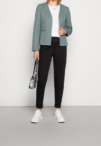 Blazer bleu sarcelle en tissu texturé, porté sur une chemise blanche, associé à un pantalon noir et des baskets blanches. Petit sac à motifs tenu à la main.