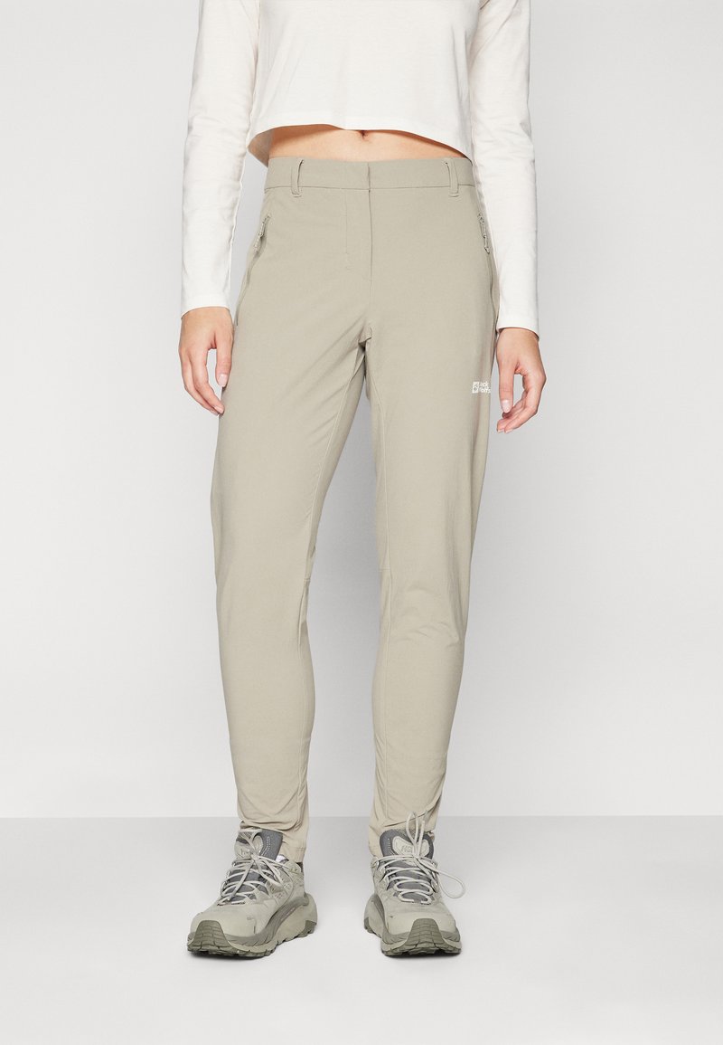Lyse beige utendørsbukser med smal passform, laget av elastisk stoff. Har glidelås-lommer og en logo nær midjeleddet.
