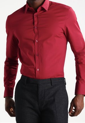 Man draagt een rood nauwsluitend overhemd met lange mouwen, ingestopt in donkergrijze nette broek, tegen een witte achtergrond.