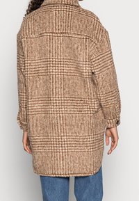 Chaqueta de cuadros marrones de gran tamaño hecha de tejido texturizado, con cuello, puños abotonados y dobladillo curvado, combinada con jeans azules.
