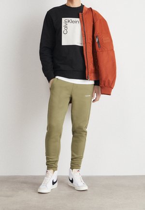 Svart sweatshirt med vit Calvin Klein-logotyp, orange bomberjacka, olivgröna joggers och vita Nike högskor.