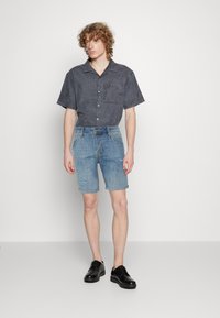 Camisa de manga corta, gris oscuro, con botones y bolsillo frontal, combinada con pantalones cortos de mezclilla azul claro con un detalle desgastado, y zapatos negros.