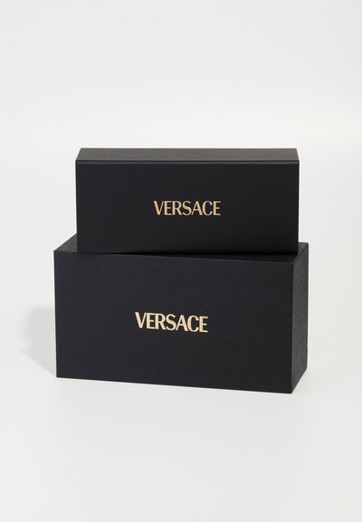 Svart teksturerte gaveesker stablet, med gull "VERSACE"-merkevare på forsiden. Den øverste esken er mindre enn den nederste.