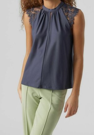 Vrouw met een mouwloze marineblauwe top met kanten details op de schouders, gecombineerd met lichtgroene, op maat gemaakte broek, staand tegen een effen achtergrond.