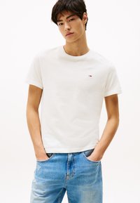 T-shirt bianco in cotone con collo rotondo, maniche corte e un piccolo logo ricamato sul petto. Indossato con jeans in denim azzurri sbiaditi.