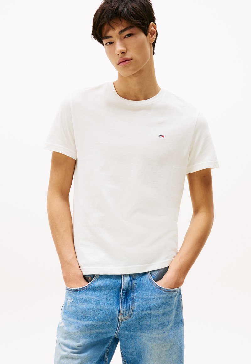 Biała bawełniana koszulka z okrągłym dekoltem, krótkimi rękawami i małym haftowanym logo na piersi. Noszona z jasnoniebieskimi dżinsami w stylu distressed.