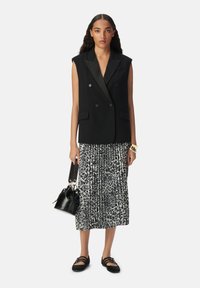 Blazer negro sin mangas con botones dobles, combinado con una falda de estampado animal en blanco y negro. La modelo sostiene un pequeño bolso negro.