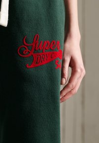 Groene stof met rode geborduurde tekst "Super Dry Co 54" naast een ontspannen hand met een lichte huidskleur.