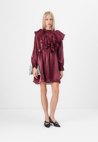 Bordeaux-rotes Seidenkleid mit Rüschendetails, langen Ärmeln und tailliertem Schnitt. Kombiniert mit schwarzen spitzen Heels und einer silbernen Kettenhandtasche.