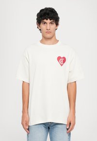Cream-colored cotton t-shirt with a loose fit, short sleeves, and a red heart graphic featuring a logo on the left chest area.