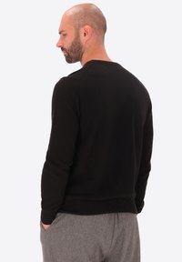 Uomo con breve barba e testa calva, vestito con un maglione nero a maniche lunghe e pantaloni grigi, con le mani in tasca, rivolto a sinistra su uno sfondo bianco.