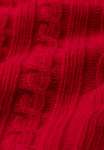 Raudona megzta medžiaga su raukšlėtomis ir pintomis raštais, minkšta tekstūra ir stora siūlai, kuriuose atsispindi jaukus ir šiltas vaizdas.
