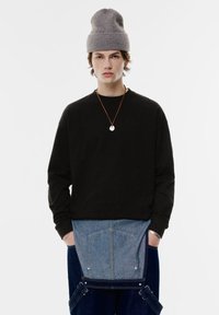 Schwarzes Langarmshirt kombiniert mit blauen Jeansoveralls, die über eine hellere Denim-Taschenverzierung verfügen. Graue Strickmütze und eine Anhänger-Halskette sind ebenfalls enthalten.