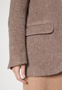 Hnědý vlněný blazer s měkkou strukturou, jednou náprsní kapsou a mírně volnými rukávy. Viditelná ruka spočívající na béžové tkanině.