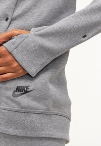 Osoba oblečená v šedé mikině Nike s otvory pro palce, ruce částečně vložené v přední kapse, na níž je vidět logo Nike.