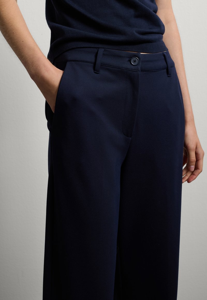 Pantaloni navy realizzati in tessuto liscio, dotati di chiusura a bottone, tasche laterali e una vestibilità rilassata con vita su misura.