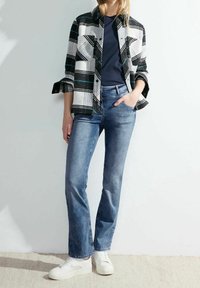 Camicia a scacchi bianca e nera con due tasche, sotto una t-shirt blu navy, abbinata a jeans a zampa d'elefante azzurri e sneakers bianche.