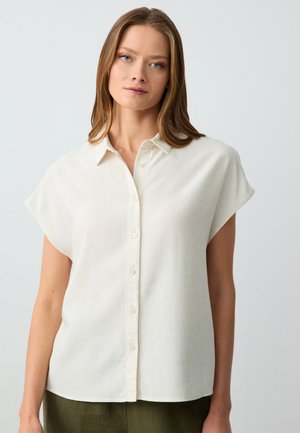 Donna con capelli lisci castano chiaro indossa una camicia bianca a maniche corte con bottoni abbinata a pantaloni verde oliva su uno sfondo neutro.