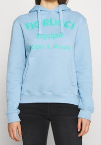 Sudadera con capucha azul claro hecha de algodón suave. Presenta texto en verde intenso y un bolsillo delantero. Puños y dobladillo acanalados que aportan estructura.