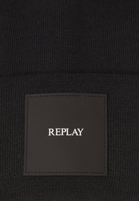 Tejido de punto negro con un parche cuadrado cosido que muestra la palabra "REPLAY" en texto blanco centrado.