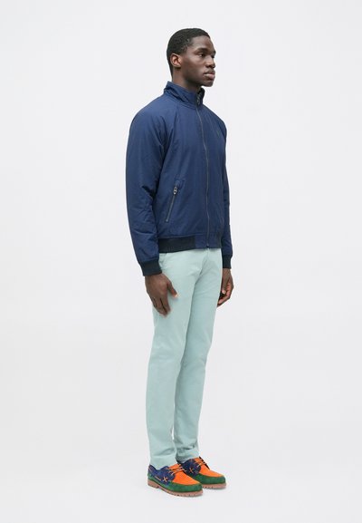 Navy bomber jacket with a stand collar, zippered front, and side pockets; paired with light mint trousers and multi-colored shoes.