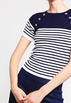 Mujer con una camiseta de manga corta a rayas horizontales en azul marino y blanco con botones plateados en los hombros y pantalones azul marino.