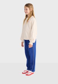 Niña joven de perfil con sudadera color crema, pantalones deportivos azules con franja blanca, calcetines blancos y chanclas rojas.