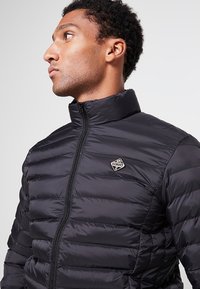 Czarna kurtka puchowa z pikowanym wzorem, wysokim kołnierzem i zamkiem błyskawicznym. Na piersi znajduje się markowe logo, a materiał ma gładką fakturę.