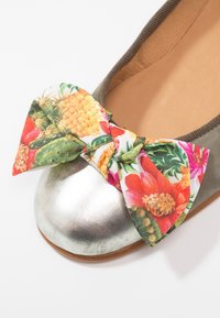 Chaussure ballerine plate argentée avec un liseré marron et un nœud en tissu coloré orné de motifs floraux et d'ananas sur le bout.