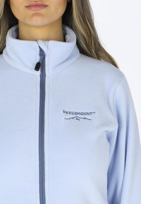 Ljusblå fleecejacka med hög krage, framdragkedja och broderad logotyp "SWEDEMOUNT" i mörkare blå. Mjuk textur och minimalistisk design.