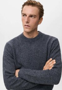 Giovane uomo con capelli castano chiaro che indossa un maglione in maglia grigio scuro, braccia incrociate, che guarda a sinistra su uno sfondo chiaro e semplice.
