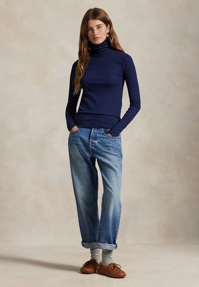 Kobieta stoi z rękami w kieszeniach, ubrana w granatowy golf, luźne niebieskie dżinsy z podwiniętymi nogawkami, szare skarpety i brązowe mokasyny.