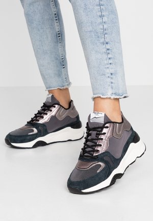 Osoba nosząca jasnoniebieskie dżinsy z postrzępionym wykończeniem i czarno-szaro-metaliczne masywne sneakersy stojąca na jasnej podłodze.