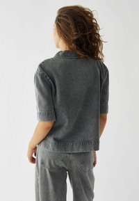 Top in denim grigio con una vestibilità comoda, maniche corte e colletto classico. Presenta cuciture visibili e una consistenza liscia, abbinato a pantaloni coordinati.