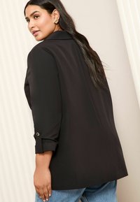 Melnā strukturētā bleizerī ar iegriezumu apkakli un saritinātām piedurknēm, kas izgatavota no gludas auduma un ar viegli pielāgotu piegriezumu, saskaņota ar ziliem džinsiem.