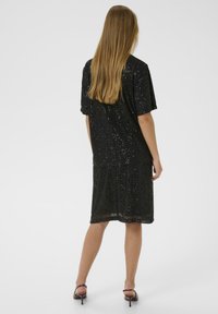 Vrouw met lang haar in een zwart pailletten jurk tot de knie en bruin met een hakken, staande met haar rug naar de camera.