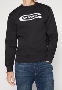 Muž v černé mikině s bílým logem "G-STAR" a modrých džínách, levá ruka v kapse a pravá ruka v uvolněné pozici.