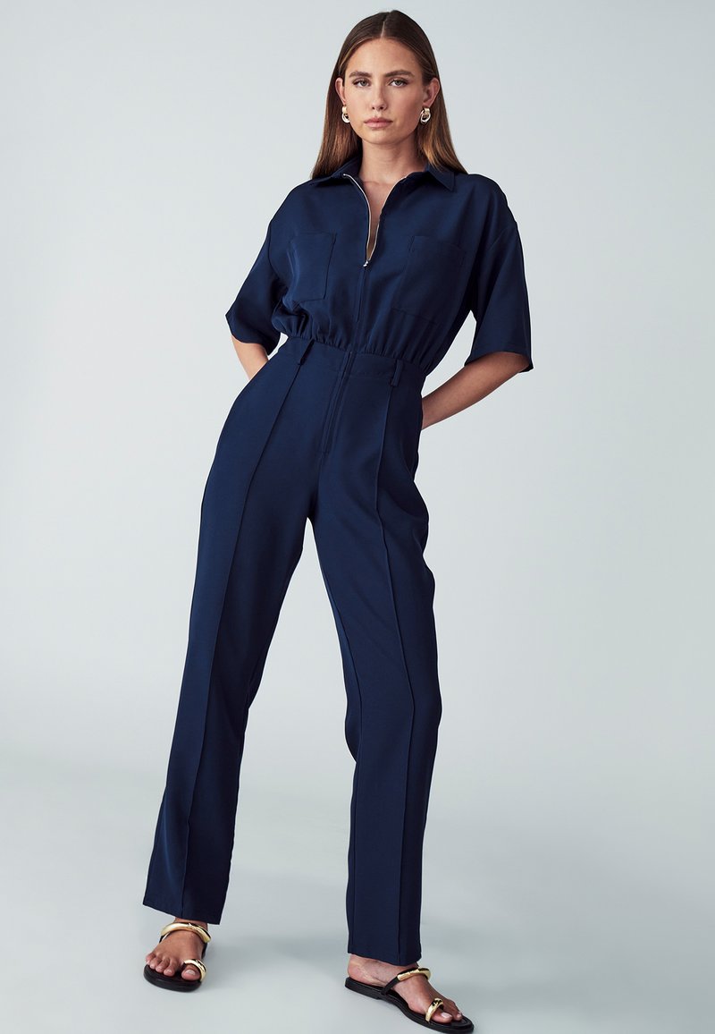 Donkerblauwe jumpsuit met korte mouwen, rits aan de voorkant en twee borstzakken. Heeft een elastische taille en een rechte, wijde pijp. Glad textuur.