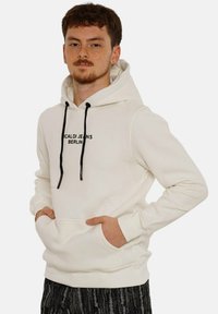 White hoodie made of soft fabric with a front pocket, featuring black drawstrings and a printed logo "CALDI JEANS BERLIN" in black.