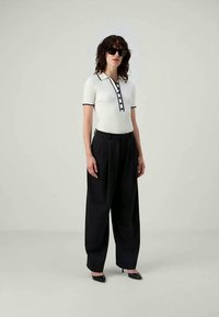 Mujer vestida con pantalones anchos negros, camiseta de polo blanca de manga corta con ribete negro, tacones puntiagudos negros y grandes gafas de sol negras, de pie.
