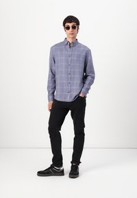 Camicia a maniche lunghe a quadri grigia con bottoni, abbinata a pantaloni neri e scarpe da ginnastica nere. Modello che indossa occhiali da sole.