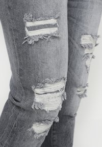 Jeans de denim gris con parches desgastados. Presentan bordes deshilachados y rayas blancas horizontales en los desgastes. Corte ajustado con una textura suave.
