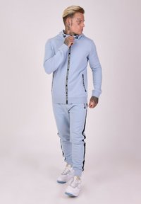 Lichtblauwe hoodie met zwarte ritsaccenten, bijpassende lichtblauwe joggers met zwarte zijstrepen, gecombineerd met witte sneakers.
