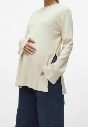 Zwangere persoon draagt een crèmekleurige top met lange mouwen en zijslitten en brede blauwe jeans, en houdt met één hand haar buik vast.