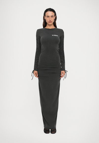 Donna in piedi che indossa un abito maxi a maniche lunghe aderente di colore grigio scuro con maniche arricciate e testo sulla parte superiore del petto.