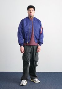 Blå bomberjakke med glidelås, maroon t-skjorte under, cargo-bukser og lyse sneakers. Enkelt bakgrunn.