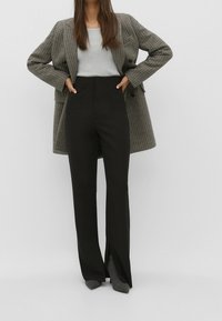 Pantalons noirs taille haute avec fentes latérales, associés à un pull côtelé gris et un blazer oversize à carreaux. Tissu texturé et design structuré.