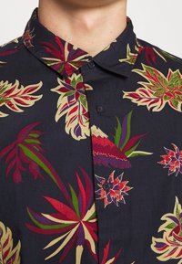Korte mouw shirt van donkerblauwe stof met levendige bloemenpatronen in rood, groen en paars. Voorzien van een traditionele kraag en knoopsluiting.