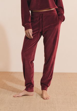 Persoon draagt bordeauxrode corduroy joggerbroek met elastische boorden en trekkoord, staat blootsvoets op een beige tapijt.